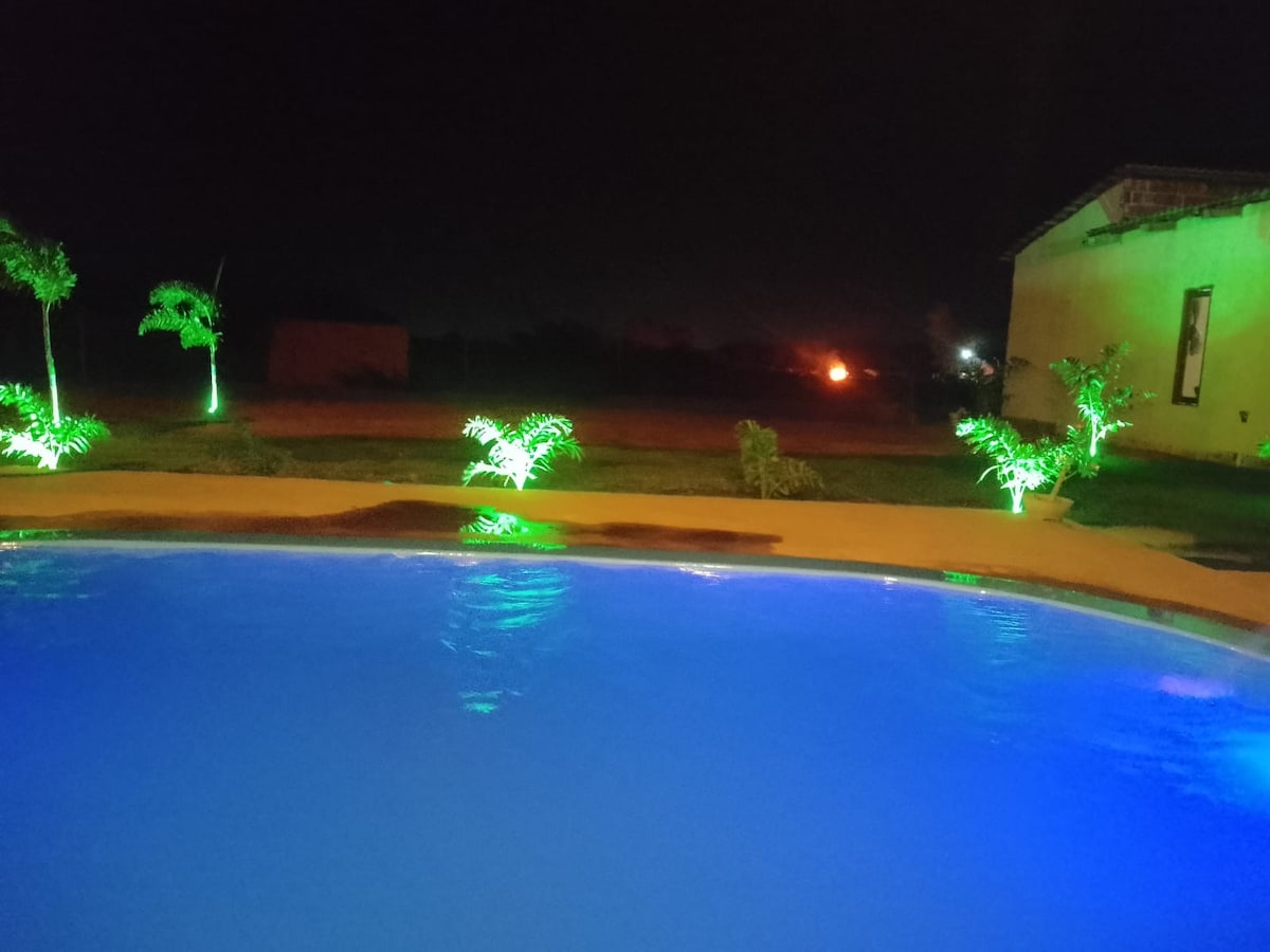 A serene pool area is illuminated by soft blue lights, reflecting a tranquil atmosphere. Lush greenery surrounds the pool, enhanced by decorative lighting that highlights the natural features. In the distance, the night sky and open landscape create a peaceful backdrop.