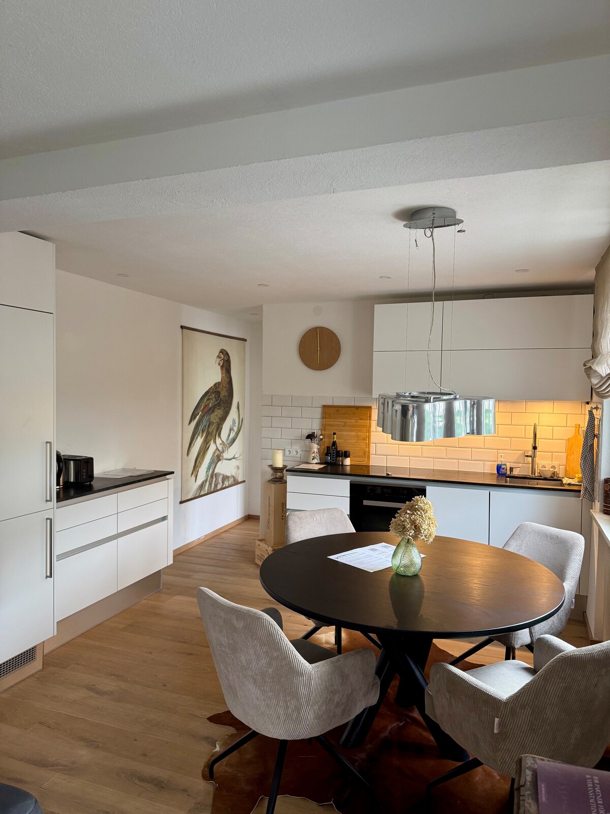 The kitchen is equipped with modern appliances including a coffee maker and a stove. A circular black dining table is surrounded by four light grey chairs. A bird mural is featured on the wall, adding a decorative touch to the space.