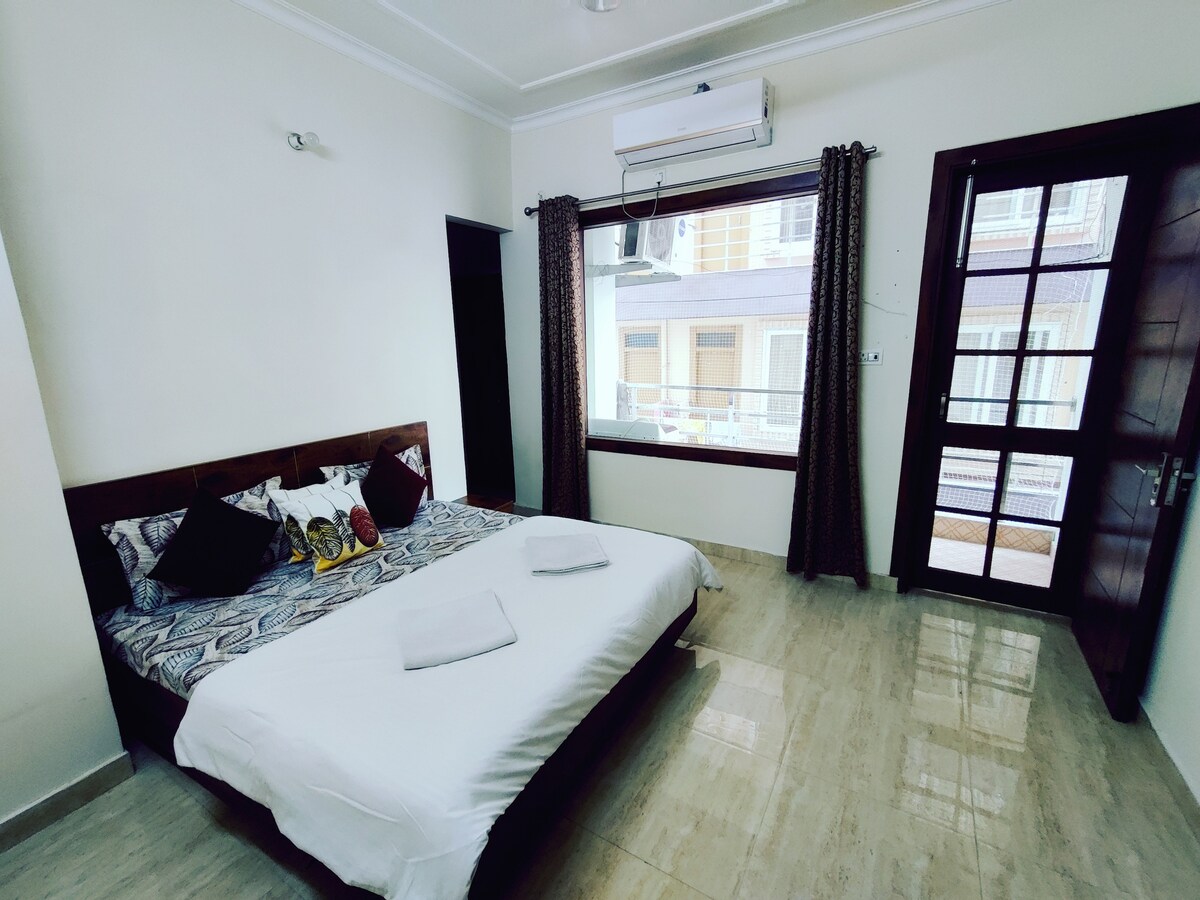 A well-furnished bedroom features a spacious bed adorned with decorative pillows. Natural light floods the space through a large window, complemented by dark curtains. The room includes a wardrobe and a door leading to a balcony, enhancing the overall layout.