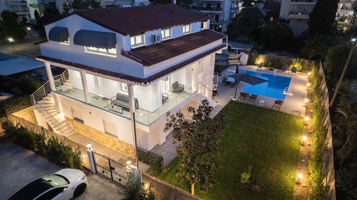A modern two-story building is set within a landscaped yard, featuring a well-lit outdoor area. A private pool and lounge chairs are positioned to the side, while surrounding greenery contributes to the space. The exterior showcases a terracotta roof and large glass windows.
