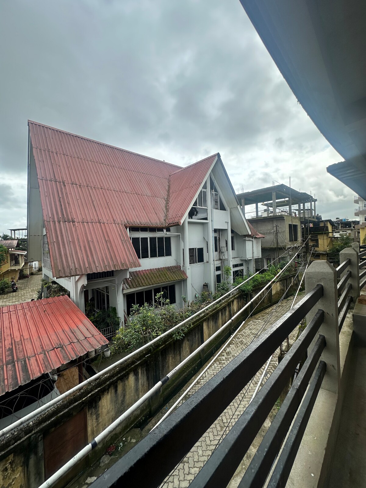 A view from a balcony features a blend of architectural styles, highlighting a building with a distinct sloped red roof and a white facade. Nearby structures including scaffolding indicate ongoing construction, while greenery offers a touch of nature amidst the urban setting.
