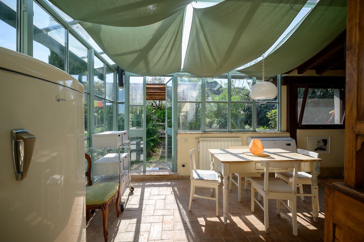 A bright dining area is framed by large windows, allowing natural light to fill the space. A light wooden table is surrounded by white chairs, with a unique pendant light hanging above it. Green fabric drapes overhead, adding a soft touch to the ambiance.