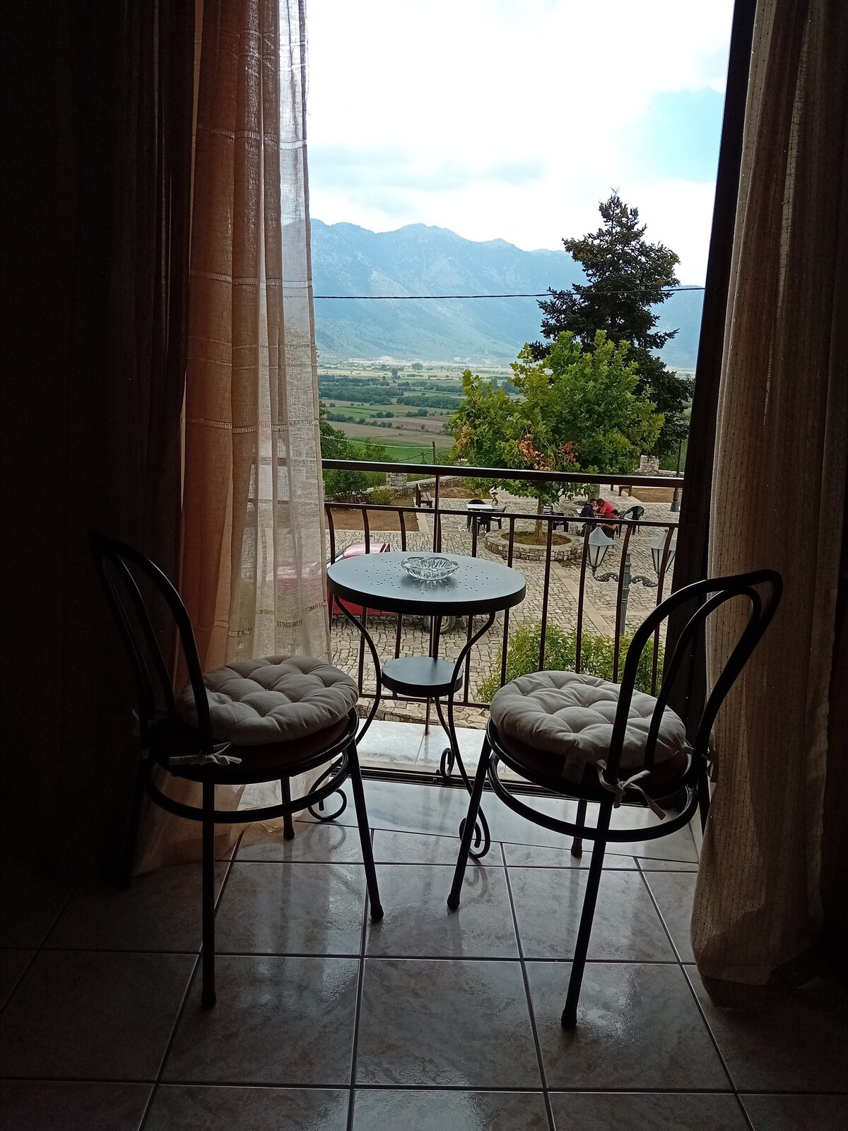 A small balcony area features two black metal chairs with cushioned seats and a round table. Sheer curtains frame the open door, allowing a view of the surrounding mountains and countryside, with greenery visible in the distance.