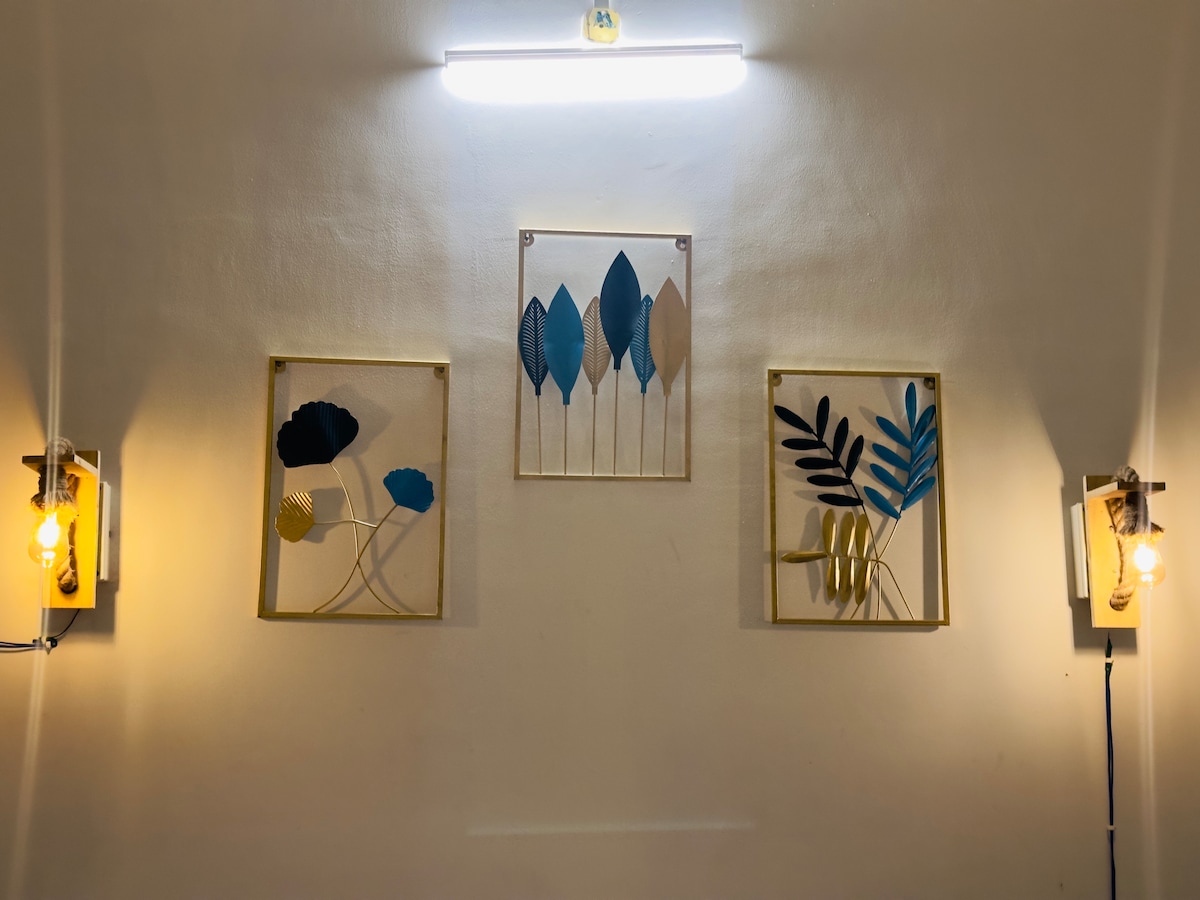 Three art pieces adorn the wall, framed in gold. Two pieces feature a combination of blue and yellow leaves, while the central piece has a delicate design of blue leaves. Soft lighting from wall sconces highlights the artwork.