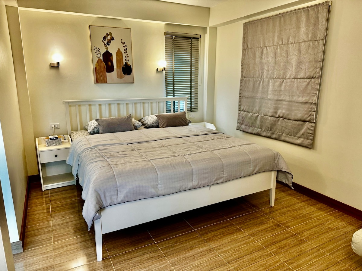A spacious bedroom features a large white bed with neutral-toned bedding and decorative pillows. Artwork hangs above the bed, complemented by soft lighting from wall sconces. Natural light enters through a window covered with a textured shade, enhancing the room's serene ambiance.