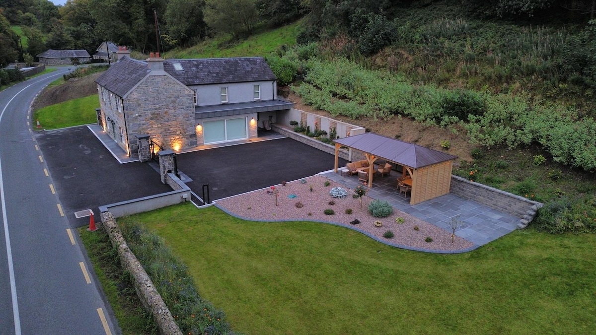 An aerial view captures Drumconnick House set amidst lush greenery, showcasing a well-maintained lawn and a stylish outdoor seating area under a wooden gazebo. The house is positioned near a quiet road, with a spacious driveway and modern stonework complementing the natural surroundings.