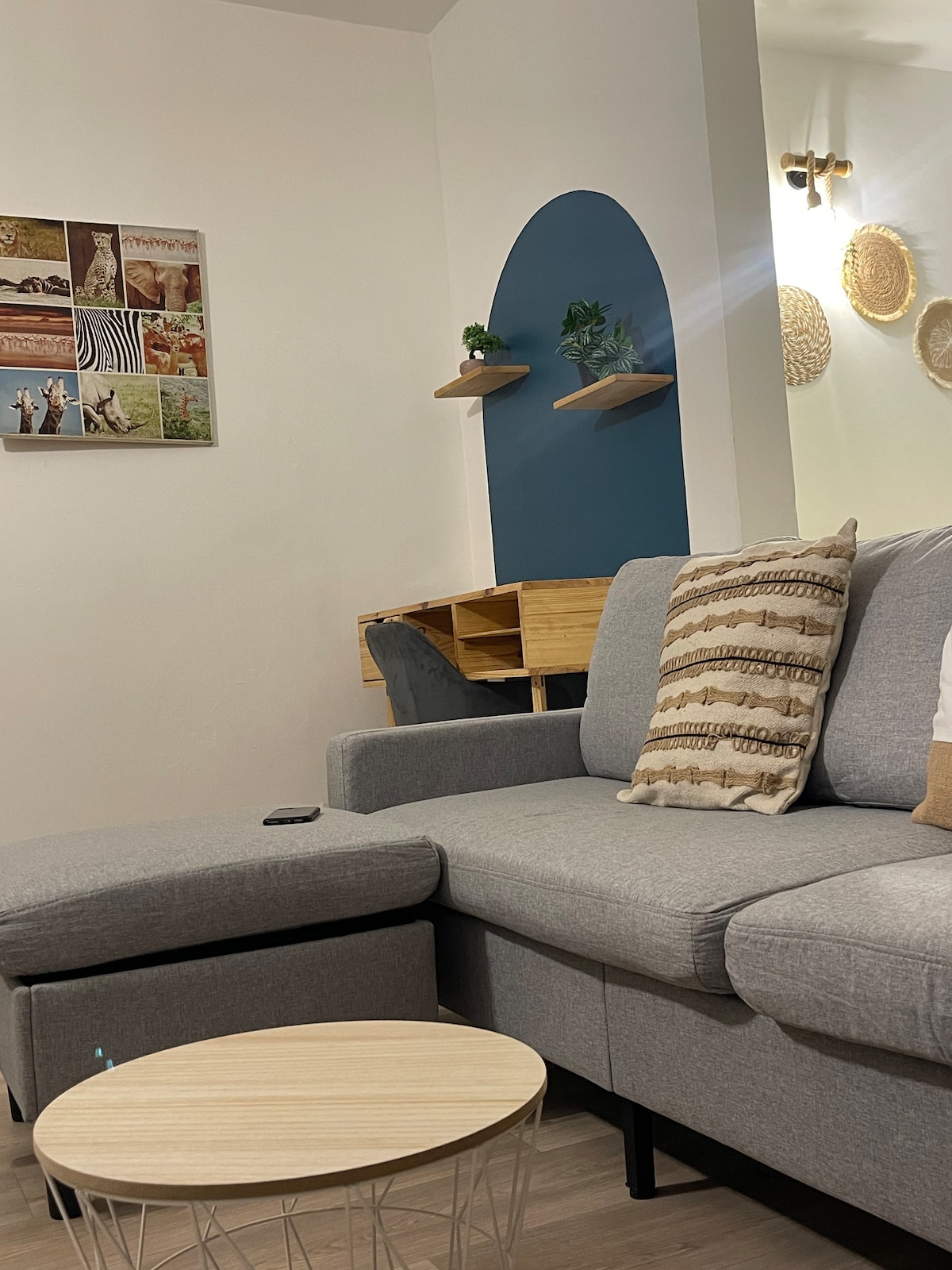 A corner of a cozy living room features a gray sectional sofa adorned with decorative pillows. A round light wood coffee table sits nearby. The wall is decorated with a framed collage of images and floating shelves with small plants. Soft lighting enhances the inviting feel.