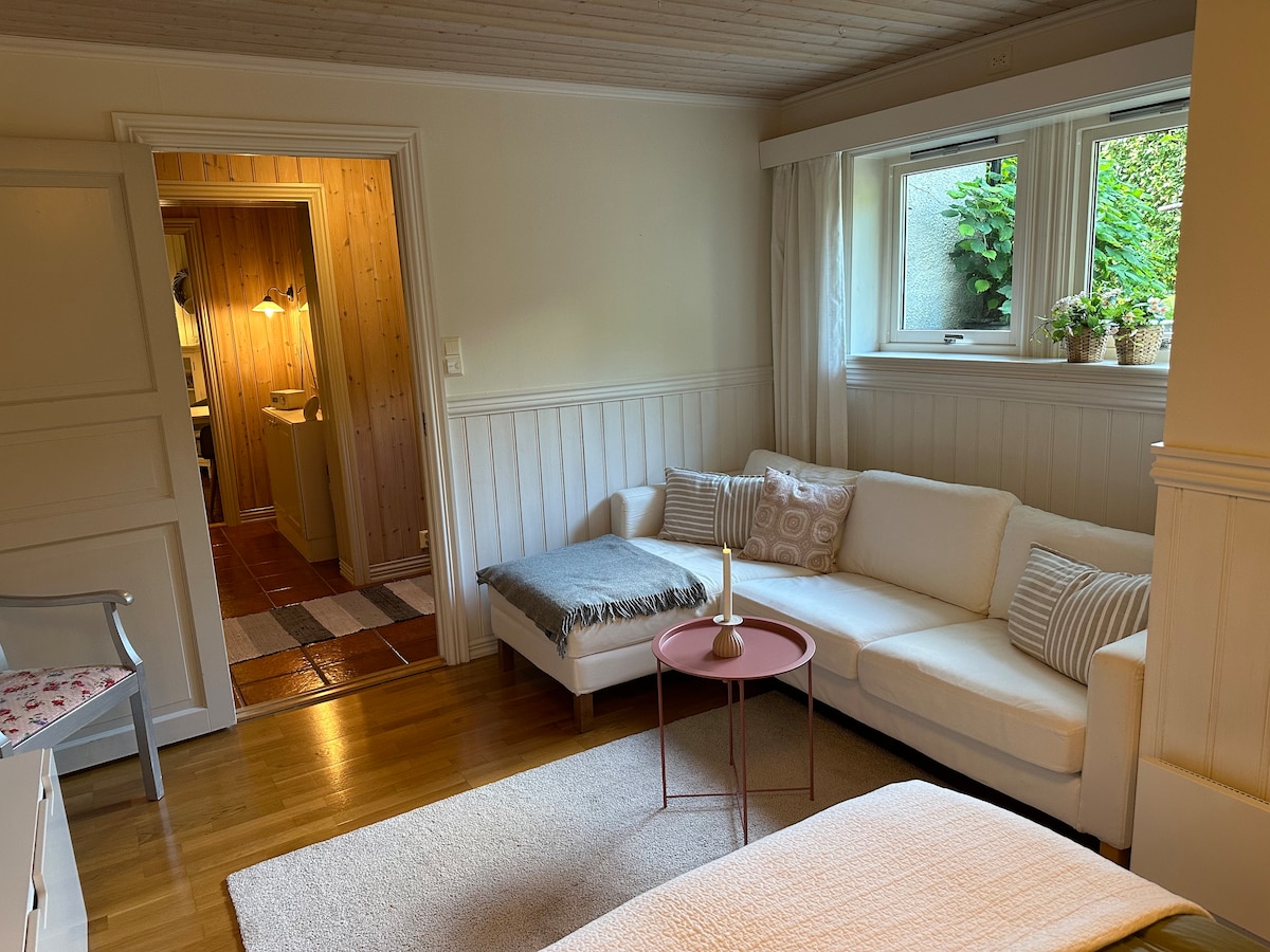 A bright living area features a white sectional sofa adorned with patterned cushions, accompanied by a small round table. Natural light enters through the window, where potted plants add a refreshing touch. A hallway leads to other rooms, enhancing the space's openness.