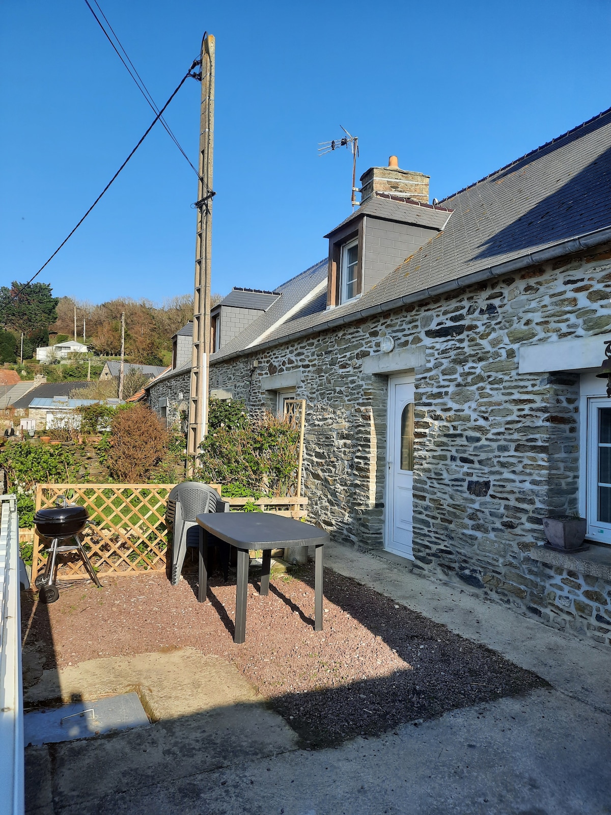 A sunlit outdoor area is presented, featuring a stone façade of the house. A dark table is positioned on gravel, accompanied by a chair and a small grill nearby. Greenery and low fencing are visible in the background, with a clear blue sky above.