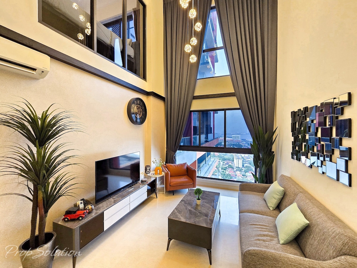 A spacious living area is highlighted by large windows providing significant natural light. A comfortable sofa sits opposite a modern TV unit. Decorative plants add a touch of greenery. Unique wall art and soft lighting create a welcoming atmosphere in this well-appointed space.