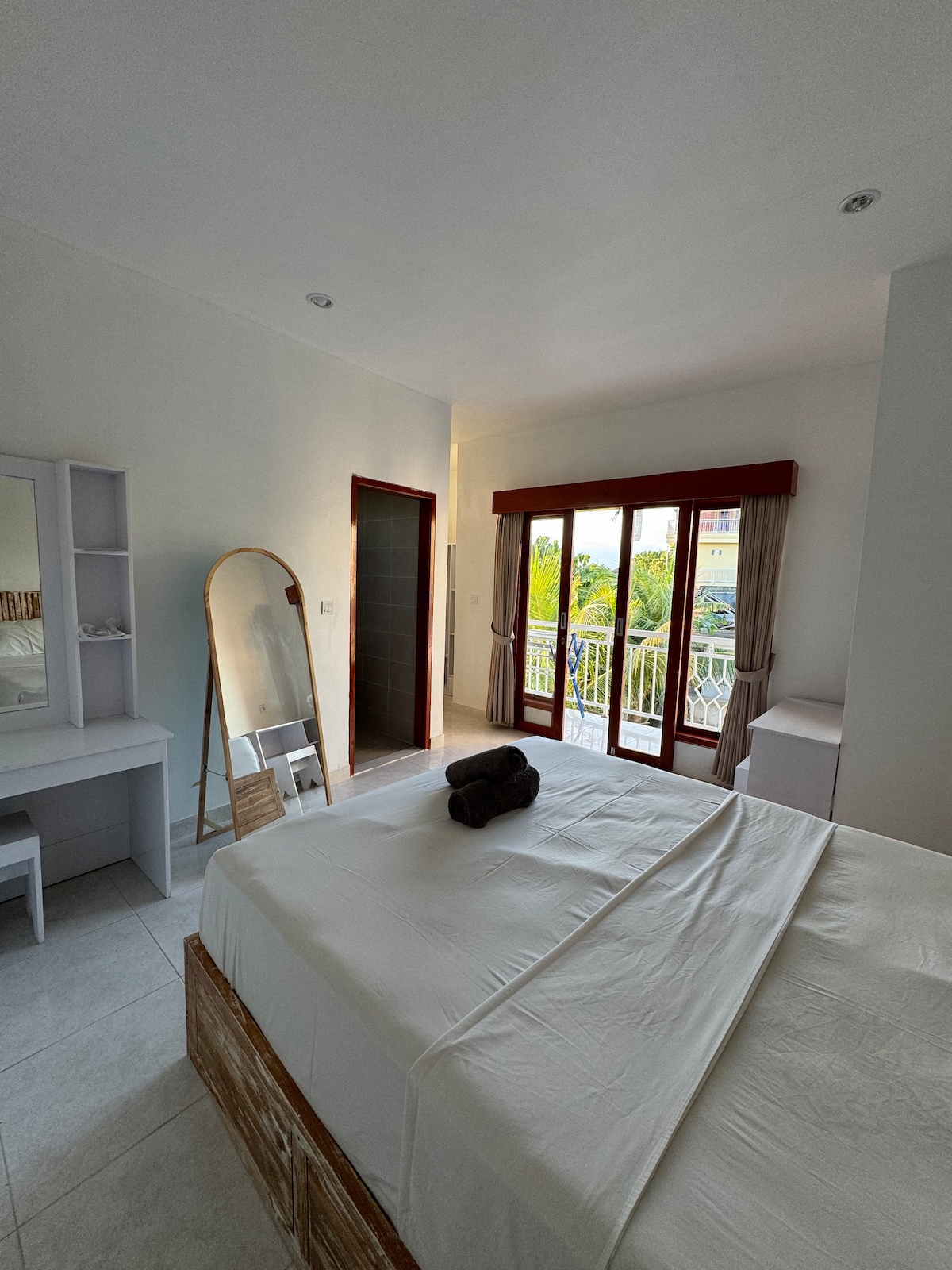 A bright bedroom features large glass doors opening to a balcony, allowing natural light to fill the space. A spacious bed is neatly made with a rolled towel placed on top. A dresser and a full-length mirror complement the minimalist decor against the light-colored walls.