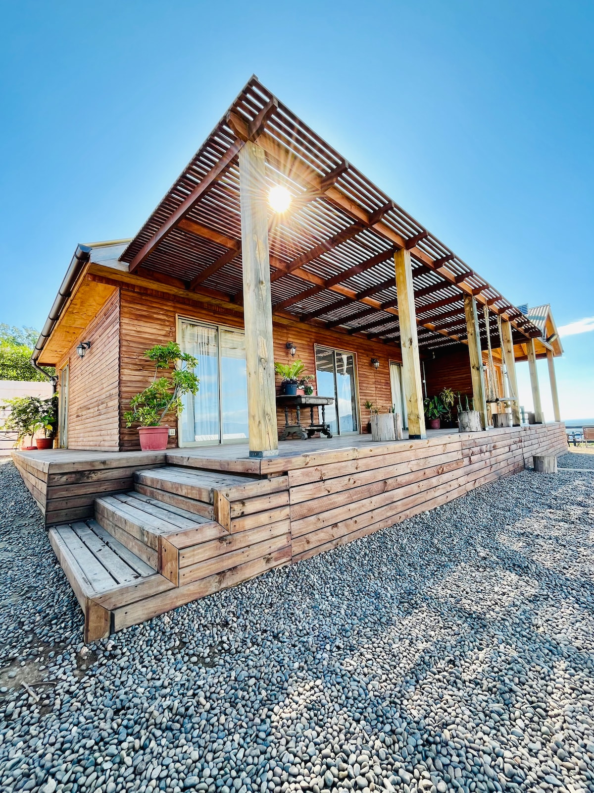 The cabin is showcased from an outdoor perspective, featuring a wooden deck with steps leading to a gravel area. Large windows are visible, inviting natural light into the interior. A roof structure provides shade over the deck, enhancing the exterior's natural appeal.