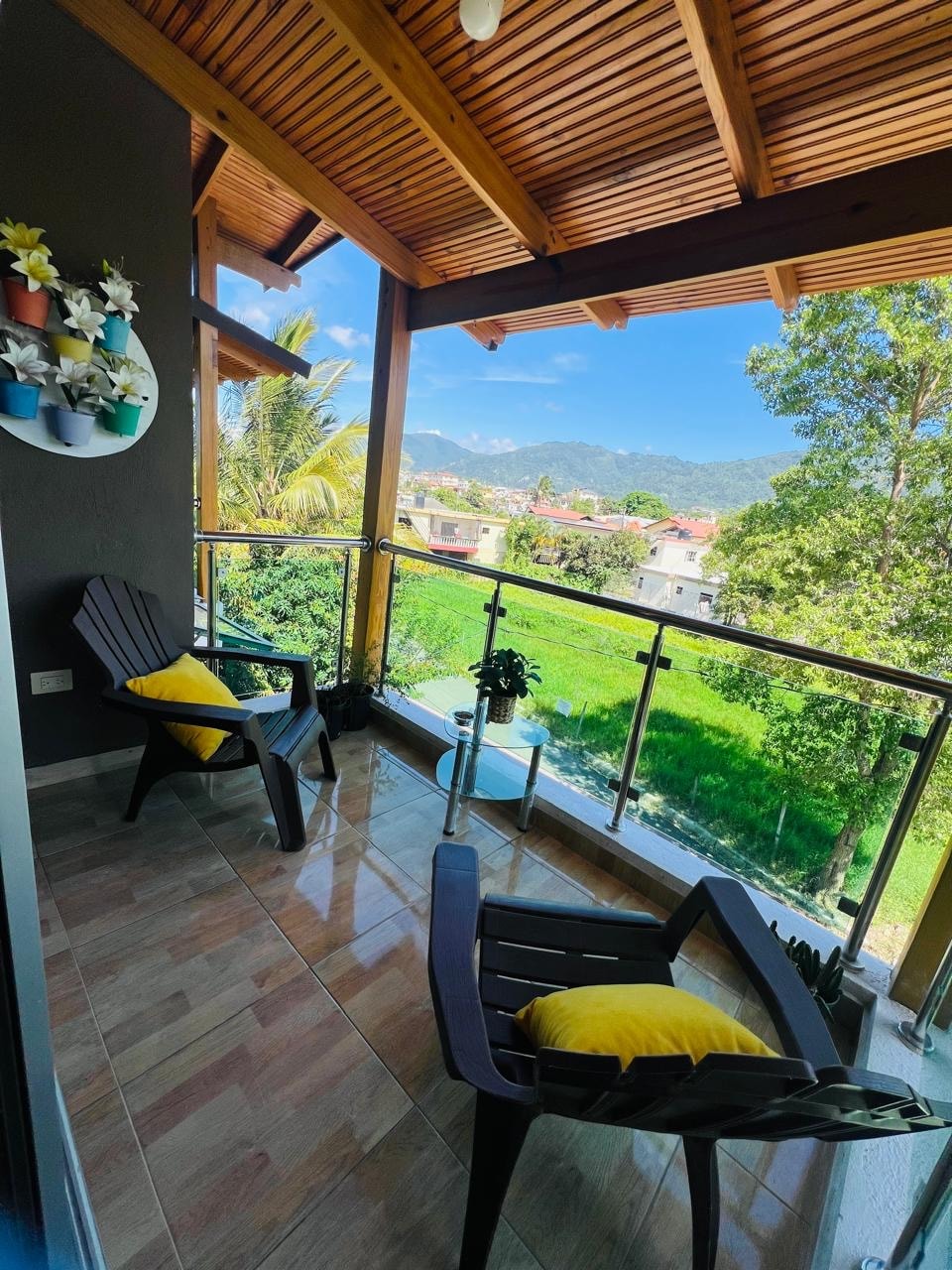 A covered balcony is presented, featuring two black chairs with yellow cushions. A small potted plant sits on the floor beside the chairs. The view includes lush greenery and distant mountains under a blue sky, framed by a wooden ceiling and railings.