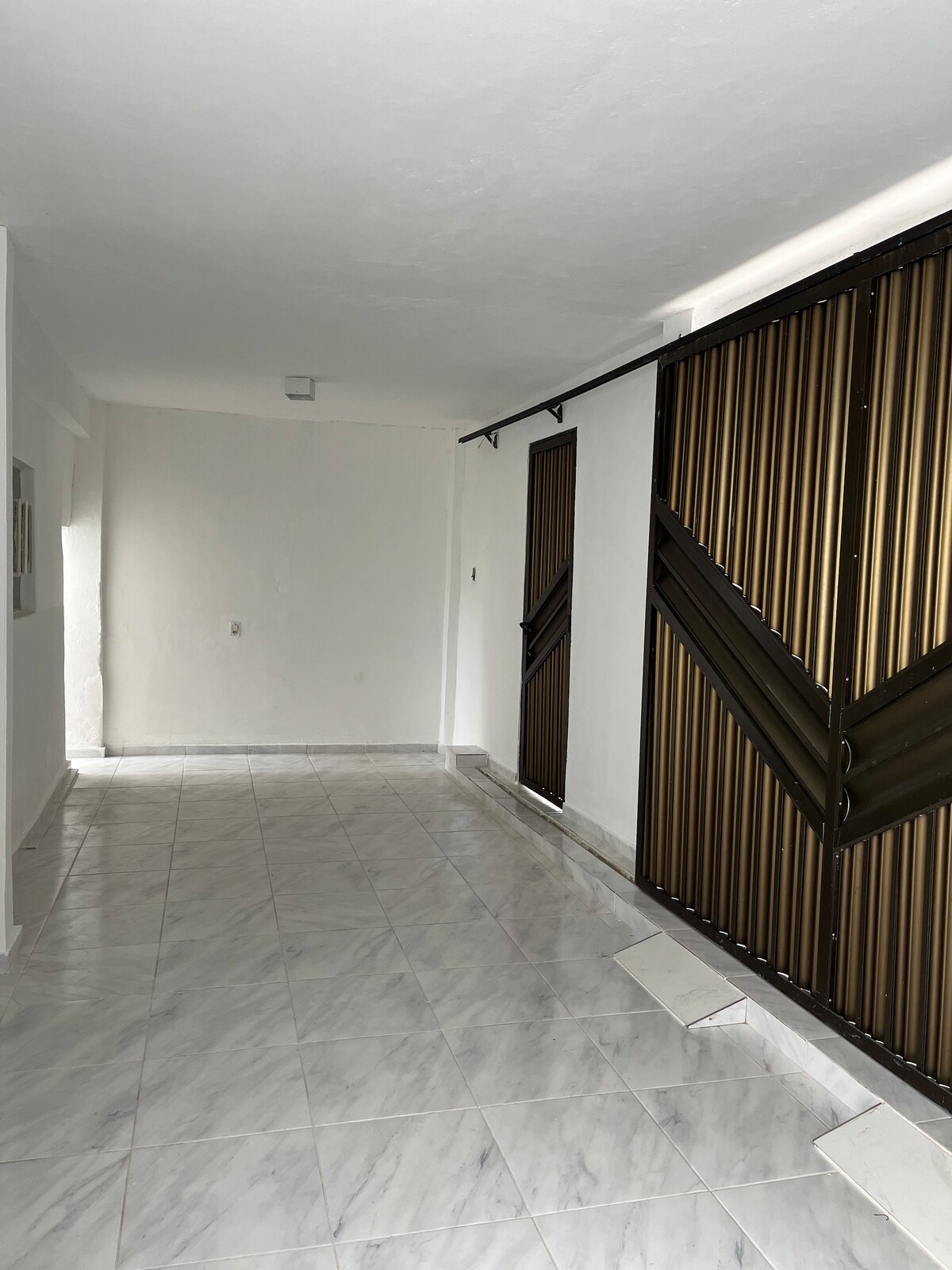 A spacious hallway features polished marble flooring, enhanced by bright, neutral walls. Two sets of large metallic doors with a distinct design are visible, allowing for ample natural light to enter the area.