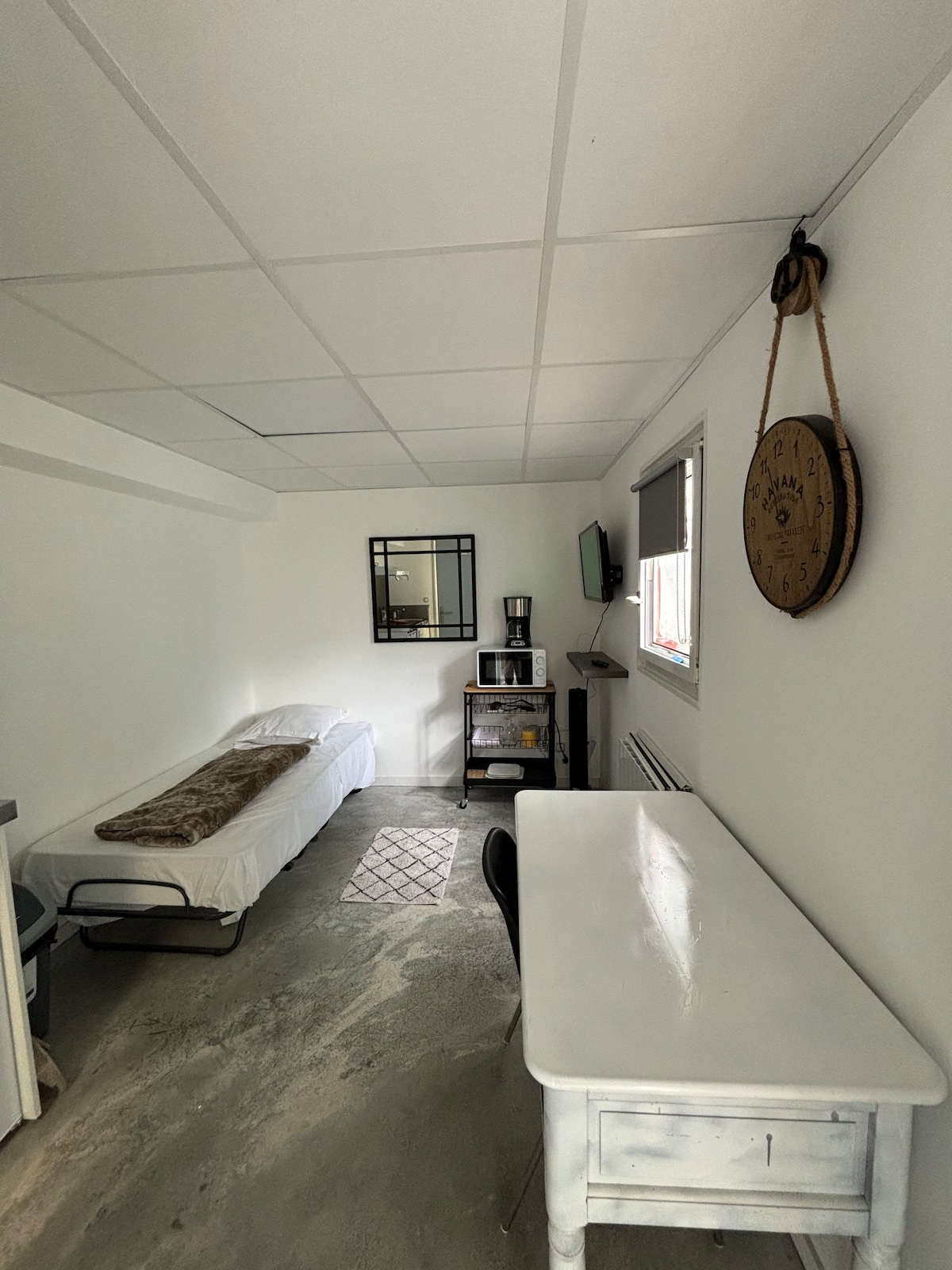 A small studio space features a single bed covered with a light-colored blanket. A desk with a chair and a small coffee station are present. A wall mirror and a clock are mounted above the desk, while a patterned rug adds texture to the concrete floor.