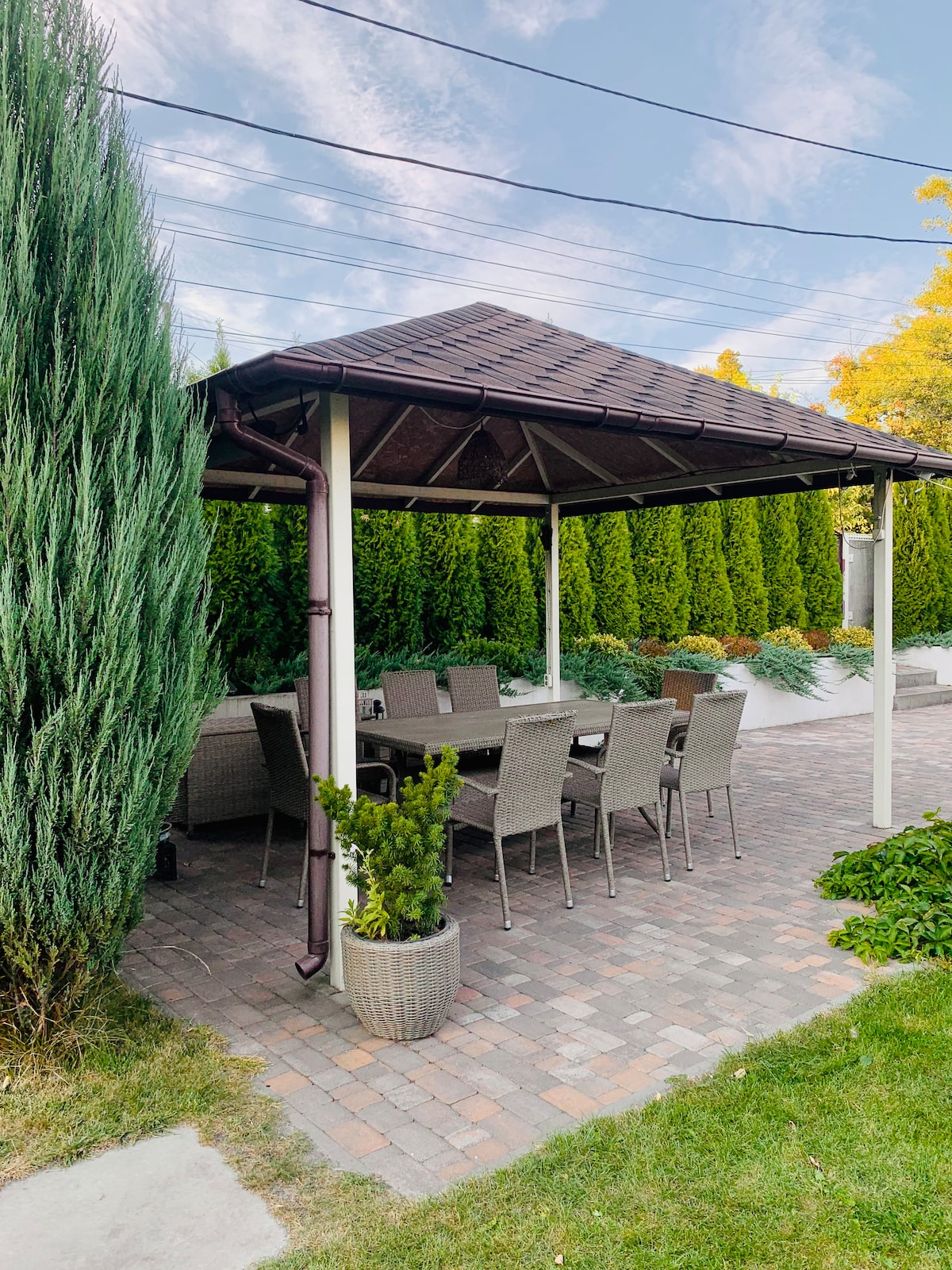 A shaded gazebo features a spacious dining area with eight gray wicker chairs arranged around a rectangular table. Lush green trees provide privacy, while neatly trimmed shrubs and flower beds add a touch of greenery to the outdoor setting.