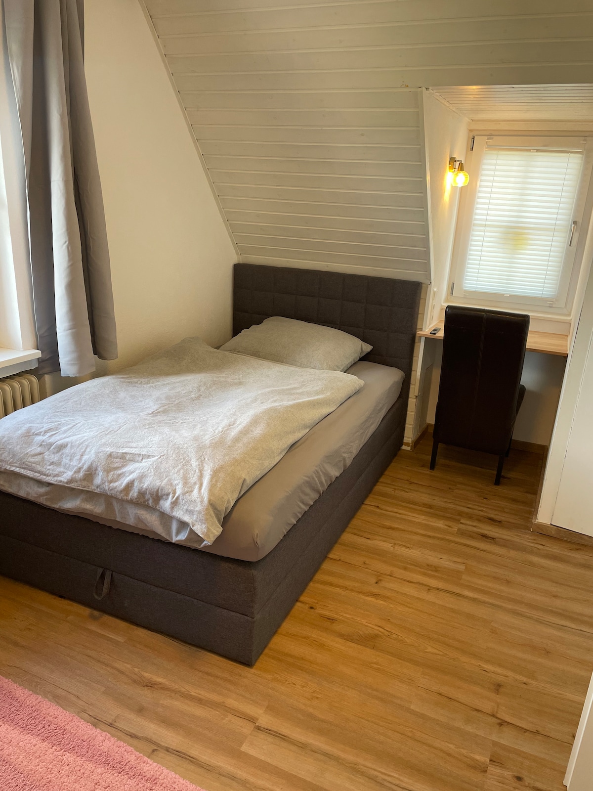 A cozy sleeping area is featured with a dark gray bed topped with soft bedding. A small desk is positioned near the window, allowing for natural light. Light wood flooring adds to the warmth of the space, complemented by light gray curtains.