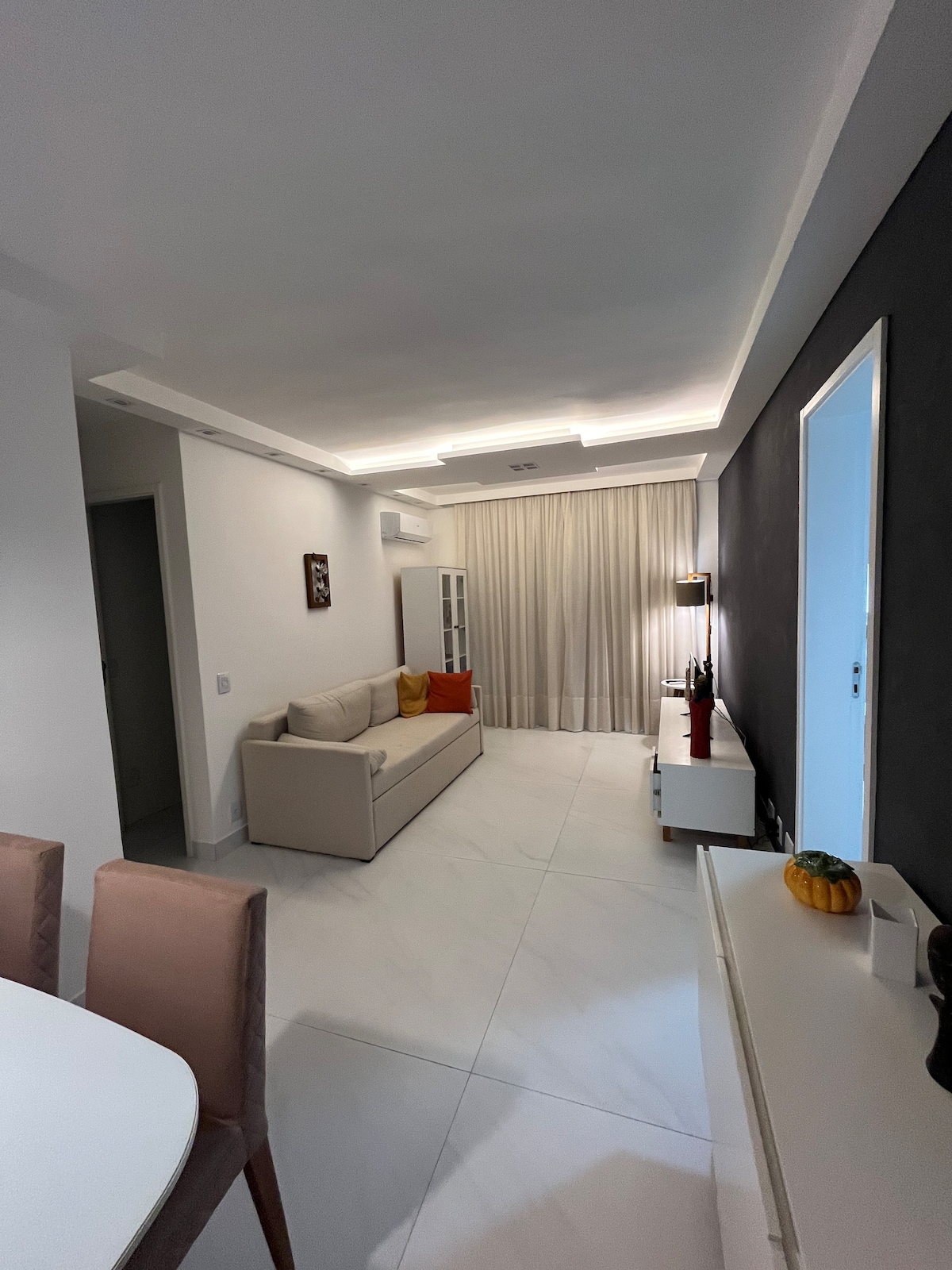 A modern living area is depicted with a neutral-toned sofa and decorative cushions. Soft lighting is present, highlighting the minimalist design. A cozy table and chairs are arranged nearby, while a cabinet and wall art add subtle character to the space. Large curtains diffuse natural light.