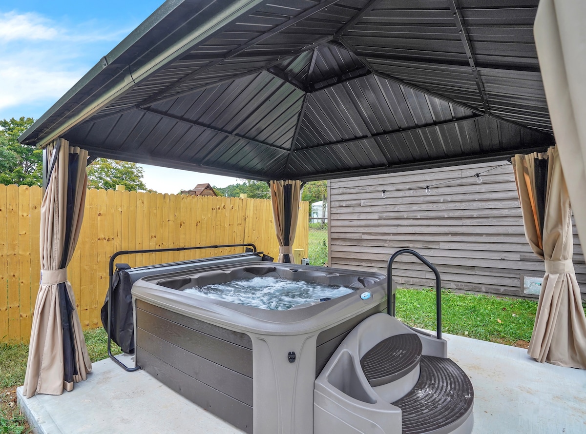 A private hot tub is positioned under a covered gazebo, surrounded by curtains for added privacy. The tub features steps for easy access and jets creating a bubbling surface. A lawn area is visible beyond the gazebo, enclosed by a wooden fence.