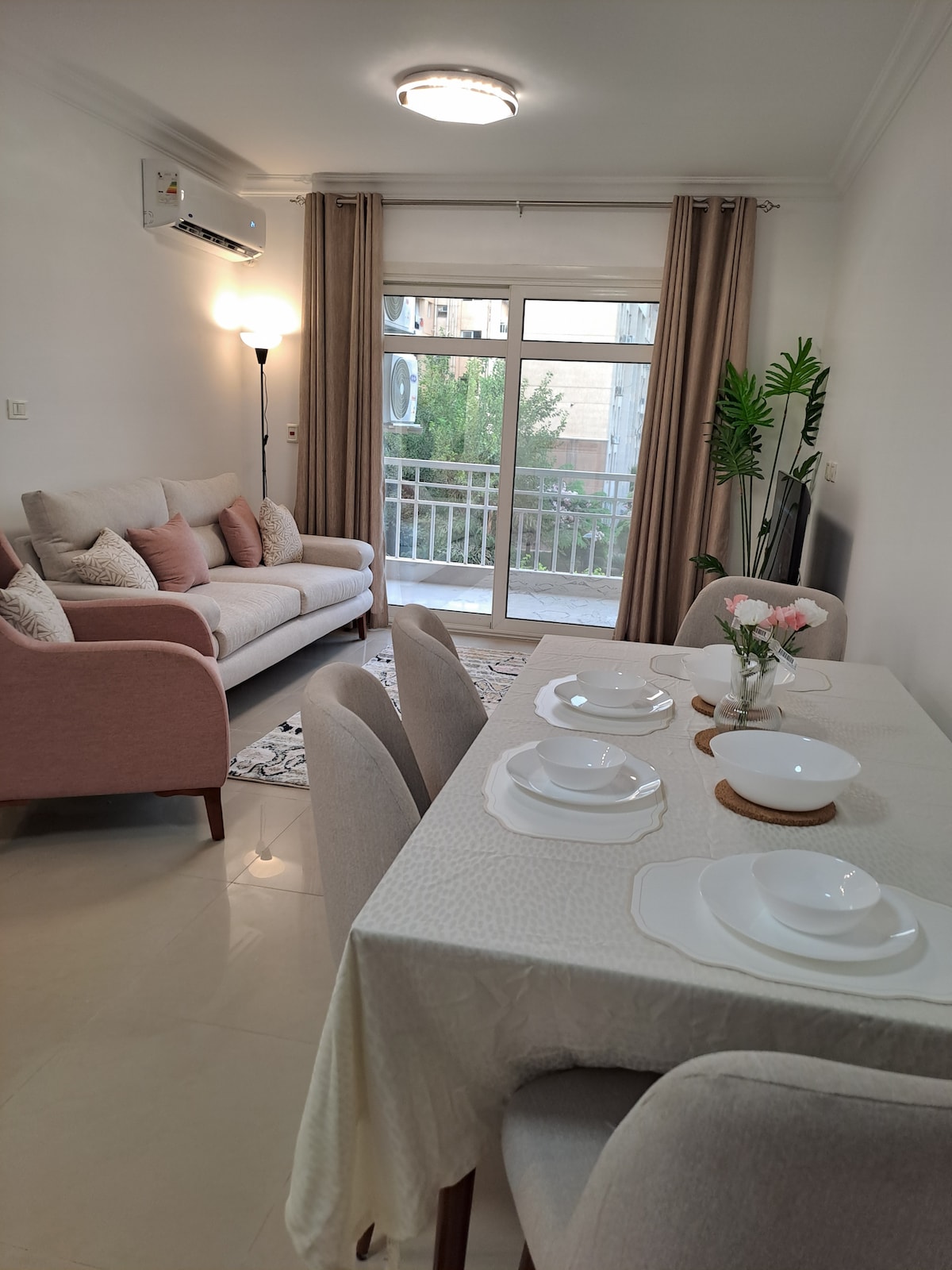 A modern living space features a soft pink sofa paired with decorative pillows, set against a backdrop of large windows. A dining table is elegantly arranged with white plates and a small floral centerpiece, creating a welcoming atmosphere enhanced by natural light.