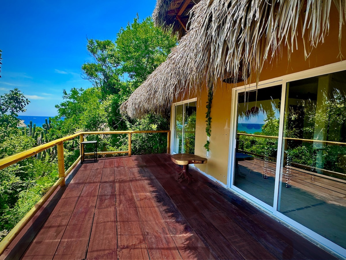 A spacious outdoor terrace is bordered by lush greenery, offering a view of the ocean in the distance. A thatched roof provides shade while large glass doors open to the interior space. A round wooden table and two chairs are positioned on the wooden floor.
