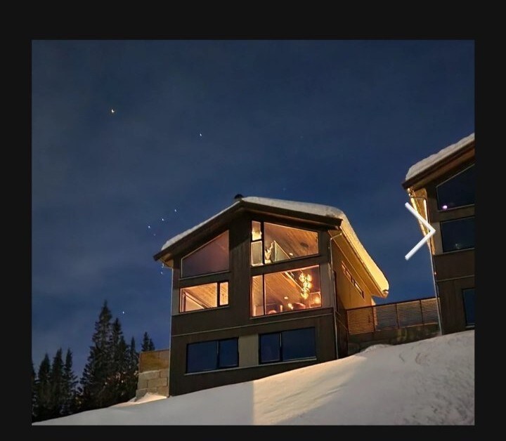 A modern cabin is situated amidst snowy surroundings, featuring multiple large windows that reflect soft indoor lighting against the twilight sky. The sloped roof and surrounding trees contribute to a peaceful mountain setting, offering a serene atmosphere for guests.