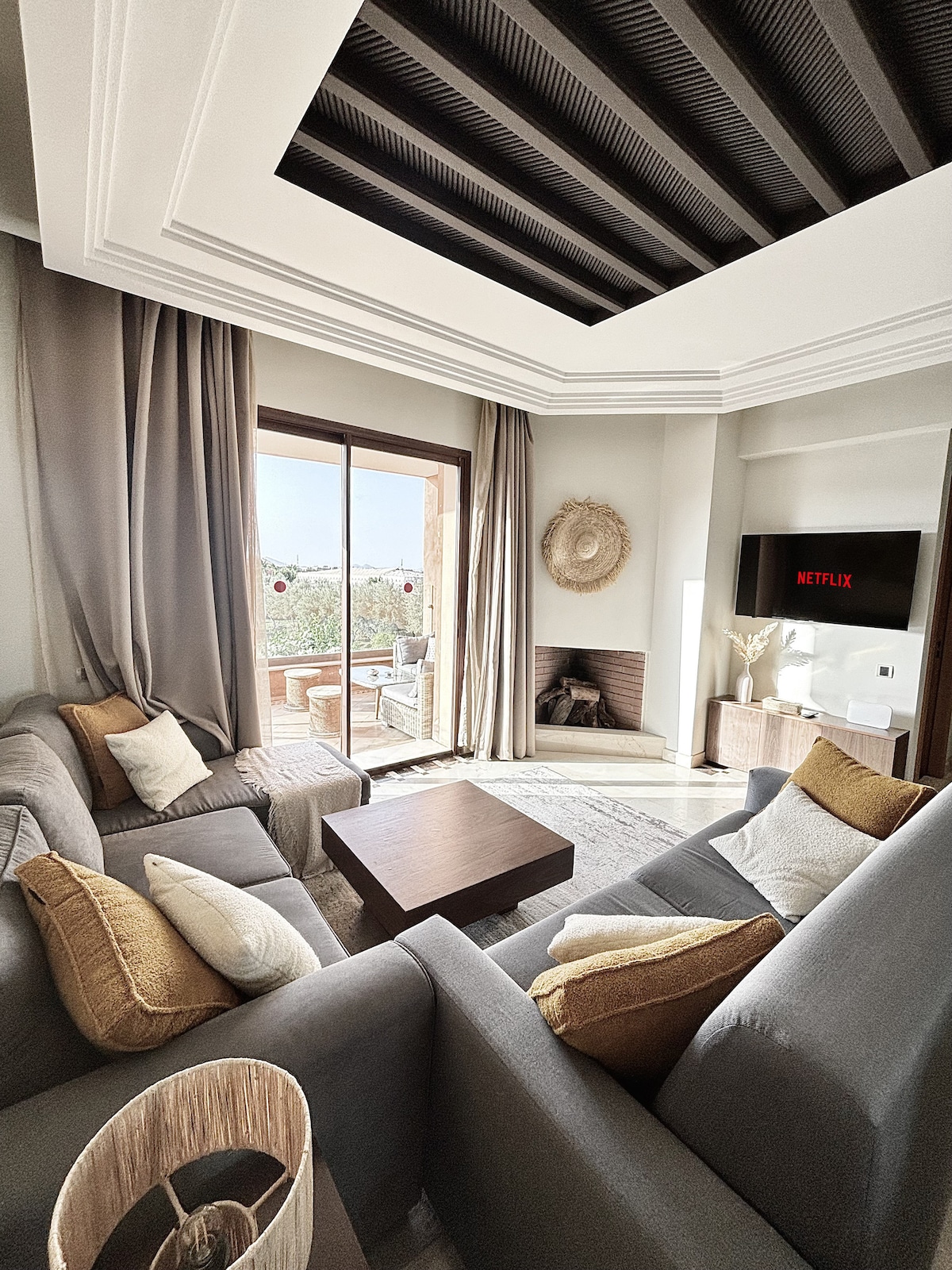 A welcoming living area features two cozy sofas with soft cushions and a wooden coffee table in the center. Natural light filters through large glass doors that open onto a balcony. A stylish fireplace and a flat-screen television are visible within the modern decor.