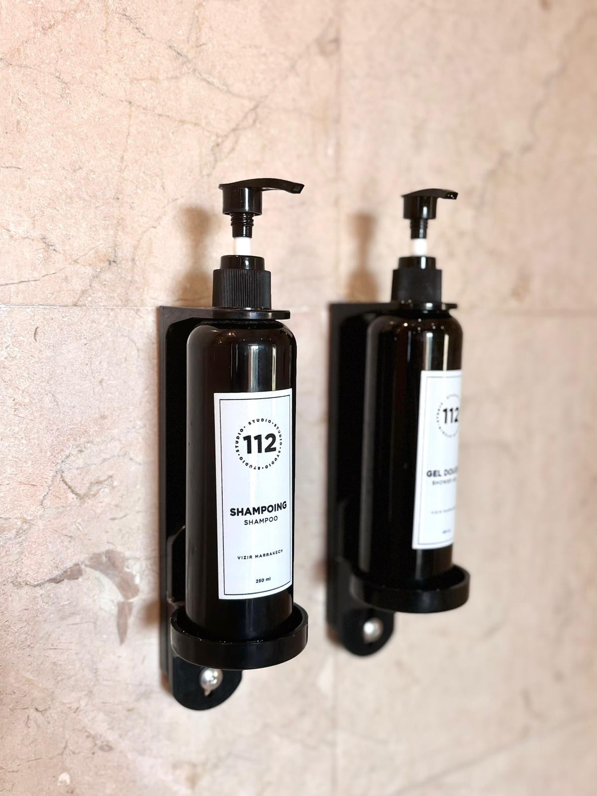 Two wall-mounted dispensers are visible, one labeled 'Shampoo' and the other 'Shower Gel.' The dispensers are sleek, featuring a dark design against a light marble backdrop. Pump tops are provided for easy dispensing.
