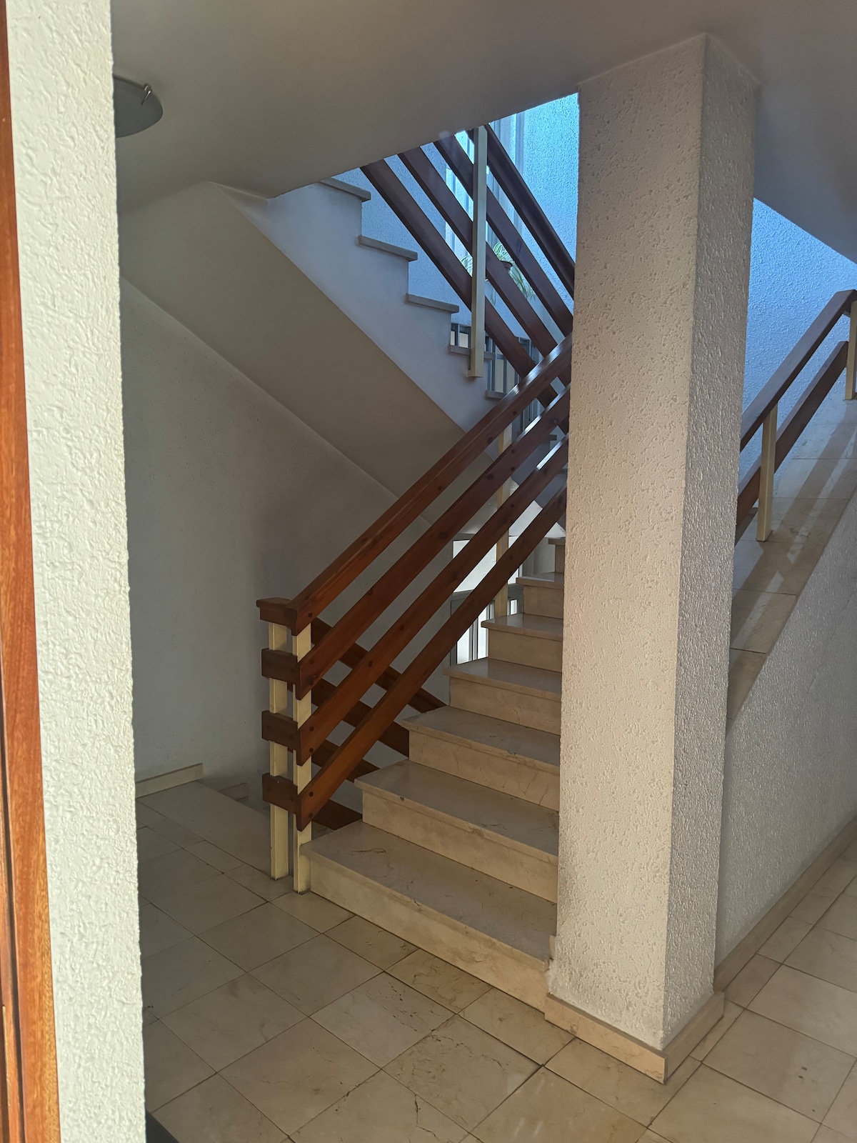 A staircase with wooden railings is seen ascending between two walls. The steps are made of light-colored stone, and natural light filters down from a window at the top. The surrounding walls are painted in a soft, neutral tone.