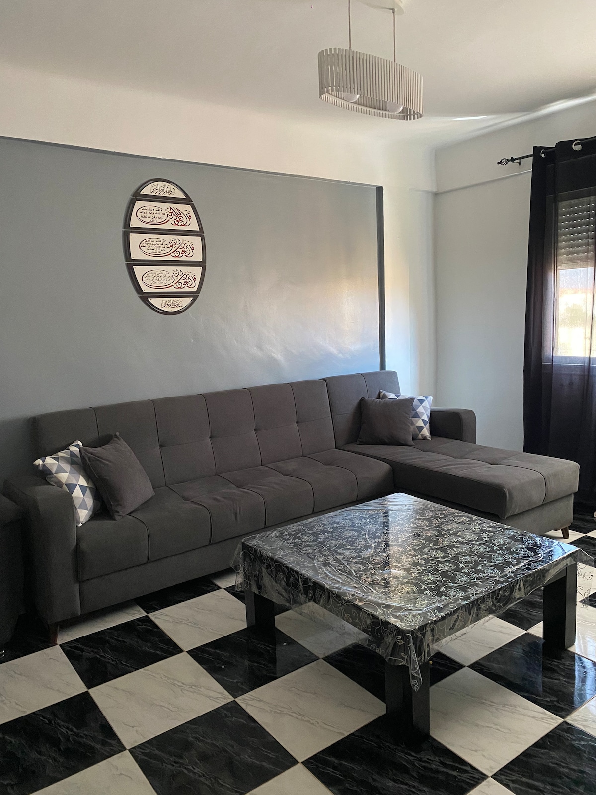 A modern living area features a grey sectional sofa complemented by decorative cushions. A glass-topped table rests atop a black and white checkered floor. Natural light enters through a window dressed with dark curtains, enhancing the space's contemporary feel.