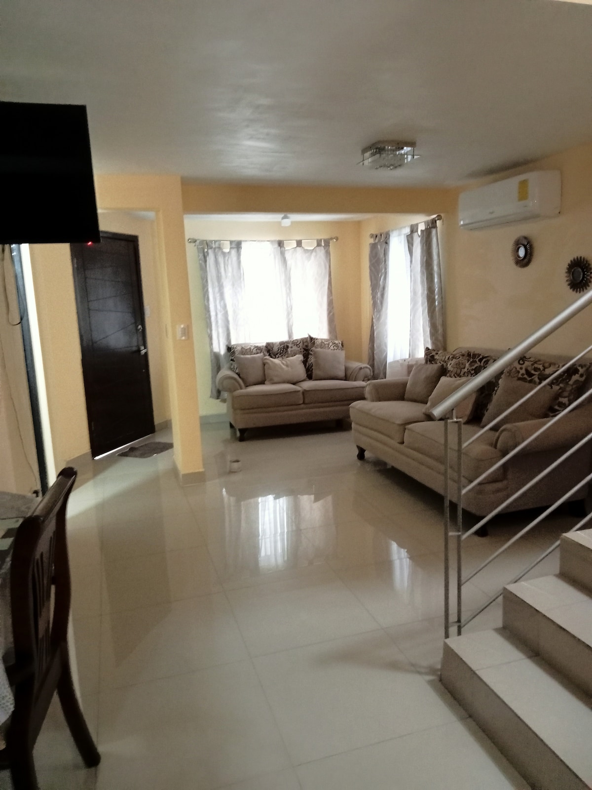 A comfortable living area is displayed with two neutral-toned sofas facing each other. Natural light enters through large windows adorned with sheer curtains. A staircase leads to an upper level, while a mounted television provides entertainment options. Soft, warm hues create a relaxing environment.