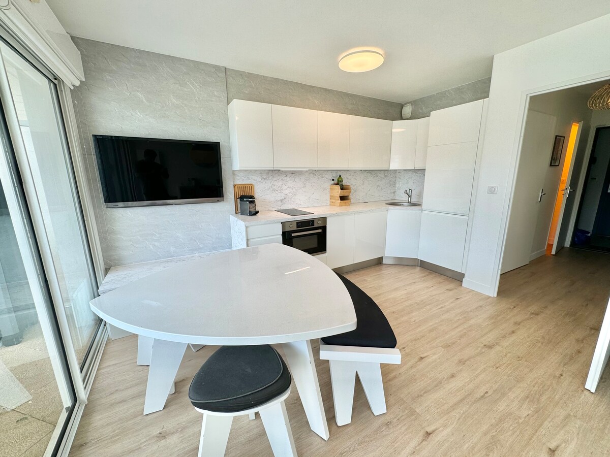 The kitchen features modern white cabinetry and a sleek countertop. A dining table with an irregular shape is paired with a curved black bench. A flat-screen TV is mounted on the wall, and a doorway leads to other areas of the apartment.