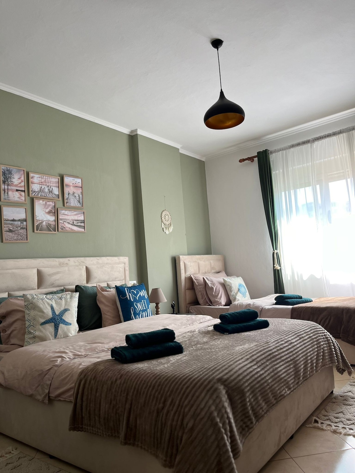 A bright bedroom is furnished with two spacious beds, each adorned with soft linens and neatly rolled towels. Green walls complement decorative wall art. A light fixture hangs from the ceiling, and natural light filters through sheer curtains, enhancing the room's comfortable ambiance.
