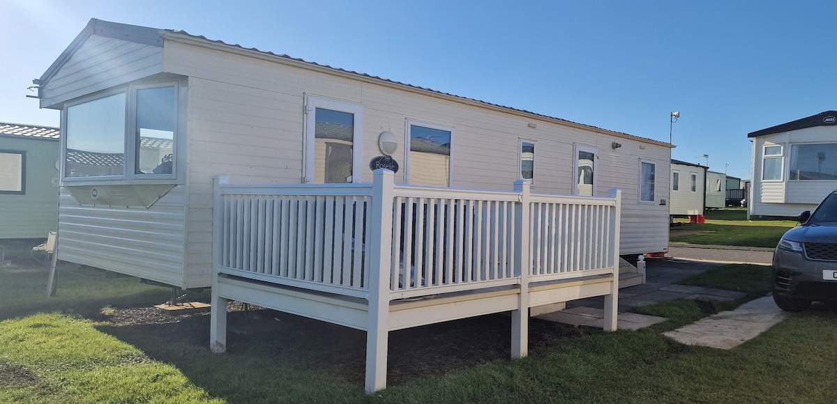 A mobile home exterior is shown, featuring light-colored siding and multiple windows allowing natural light. A spacious white deck surrounds the front entrance, providing an area for outdoor relaxation. Green grass and a clear blue sky serve as the backdrop.