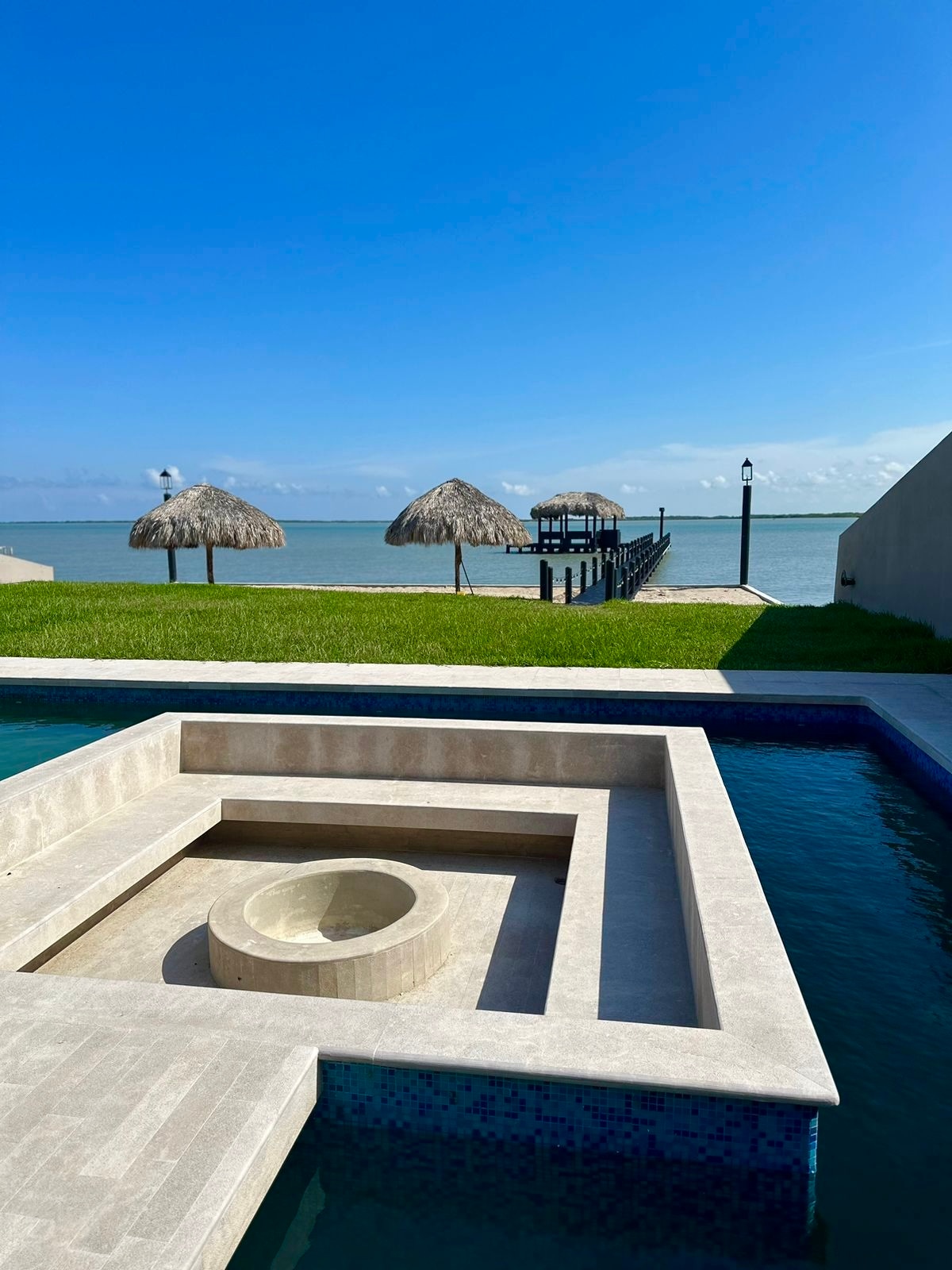 A private pool area is viewed, featuring a rectangular shape with a round inset structure at its center. Beyond the pool, thatched palapas provide shade on the grassy edge, with a wooden dock extending into the calm water under a clear blue sky.