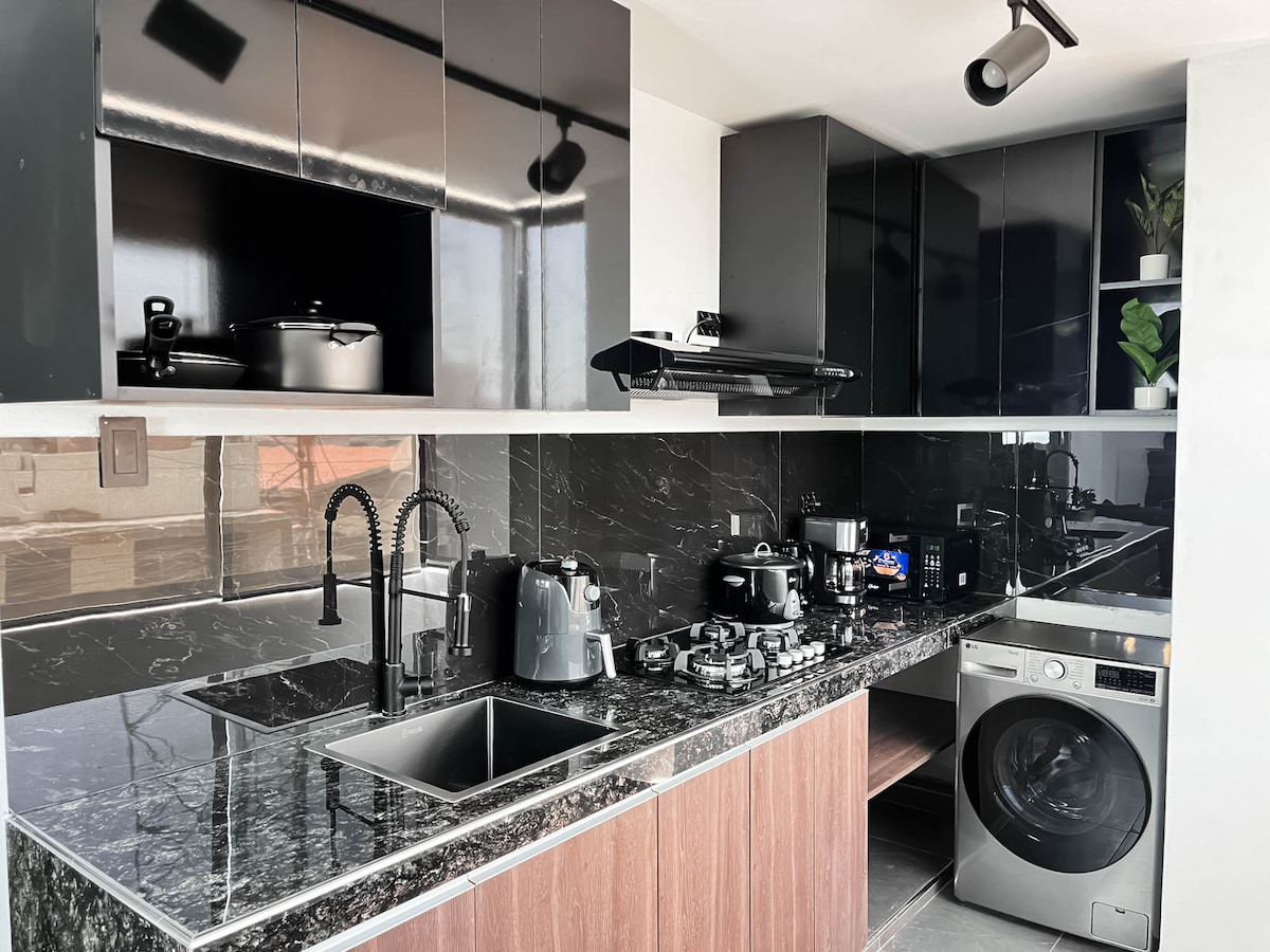 A modern kitchen showcases sleek black cabinetry and a stylish marble countertop. Kitchen appliances, including a kettle, coffee maker, and gas stove, are neatly arranged. A washing machine is integrated into the cabinetry, while large windows allow for natural light to illuminate the space.