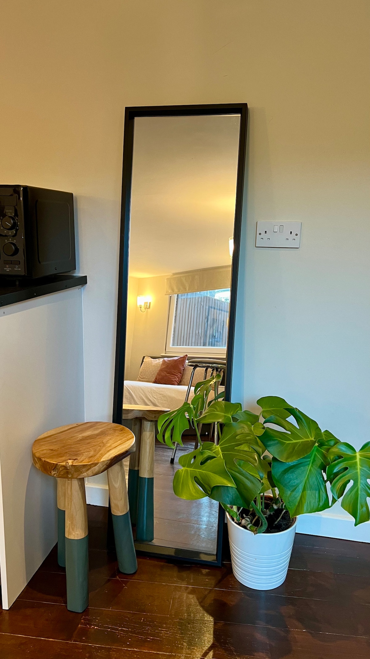 A full-length mirror is positioned against the wall beside a small wooden stool with a two-tone design. A vibrant green plant in a white pot adds a touch of nature to the space. The room is softly lit in the background, hinting at the cozy atmosphere.