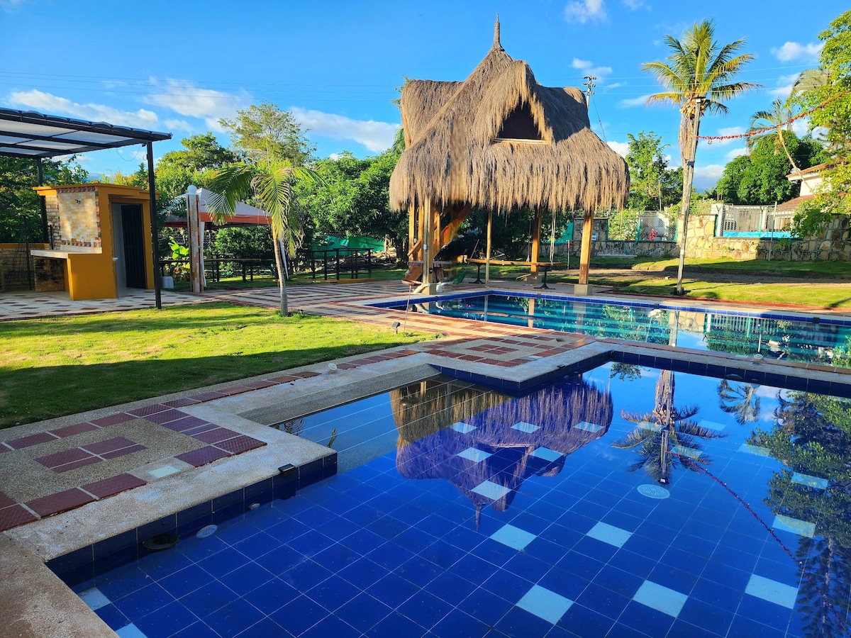 A scenic outdoor area features a sparkling blue pool surrounded by manicured grass and patterned tiles. A thatched-roof gazebo offers shade, with palm trees providing a tropical vibe. Reflections can be seen in the calm water, enhancing the serene setting.