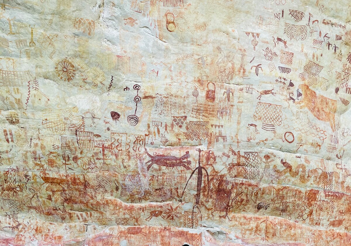 The image depicts ancient rock paintings on a stone surface, showcasing a variety of motifs including animals and geometric patterns. The colors range from earthy reds and browns to lighter shades, emphasizing the intricate designs and historical significance of the artwork.