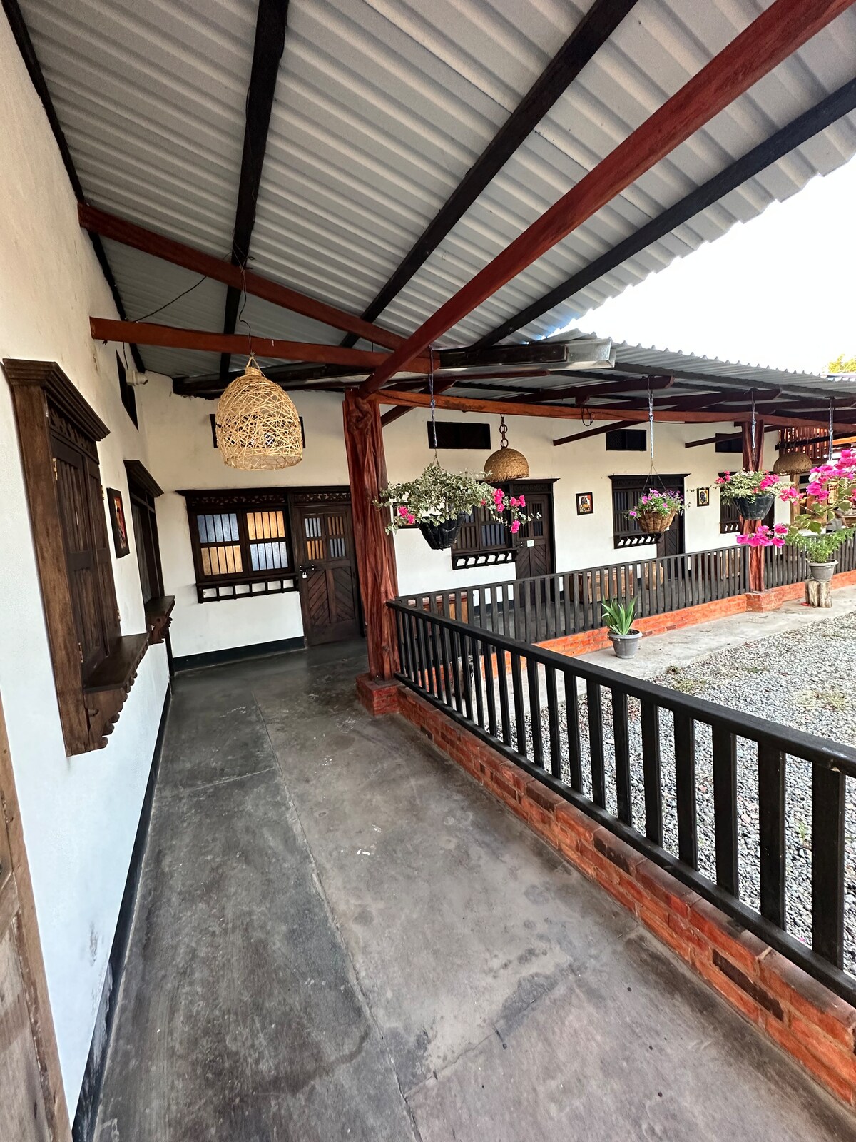 A covered walkway is presented alongside a gravel path, featuring wooden beams and railings. Decorative hanging plants are displayed, while wall art and windows adorn the white stucco walls, creating a harmonious connection with the natural surroundings.