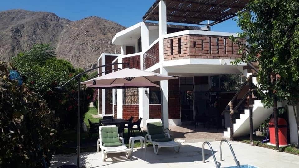 The exterior of the spacious two-story house is set against a mountainous backdrop. A shaded area features lounge chairs, while an outdoor dining table is visible. The structure has large windows, and a pool is situated in front, surrounded by greenery.
