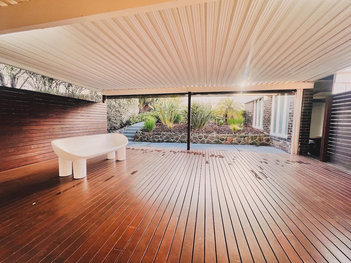 An outdoor area features wooden decking beneath a covered ceiling. A modern white tub is centered on the deck, surrounded by landscaped greenery visible in the background. Natural light filters through, offering a bright and open ambiance.