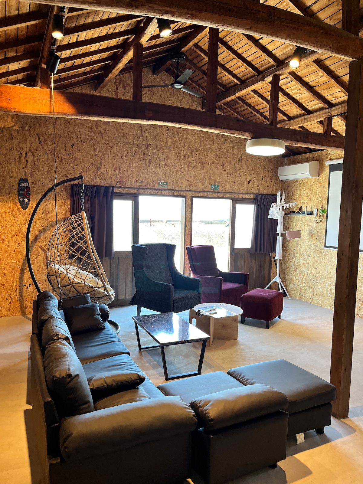 A spacious living area features a combination of seating options, including a leather sectional sofa and armchairs. Large windows invite natural light, and an overhead fan complements the rustic wooden ceiling. A glass coffee table sits centrally in the room, enhancing the open feel.