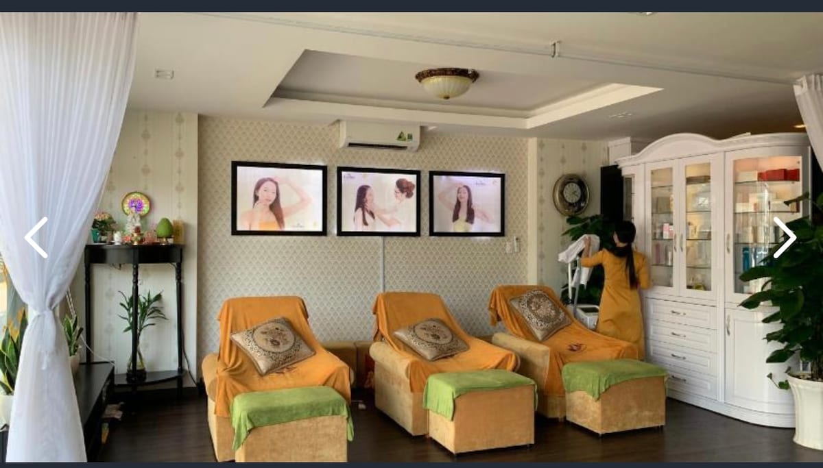 A serene spa area is presented, featuring three relaxation chairs draped with soft orange throws. Green accents add a calming touch. The wall displays framed images, contributing to a tranquil ambiance. A stylish cabinet holds decorative items, while potted plants bring a hint of nature.
