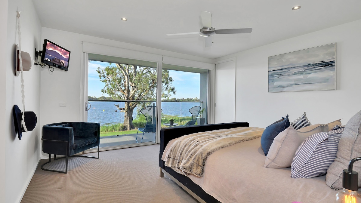 A serene bedroom features a spacious bed adorned with soft pillows, accompanied by a cozy chair and wall-mounted TV. Large sliding doors open to a balcony overlooking tranquil water views, while a tree is visible in the background, enhancing the calming environment.