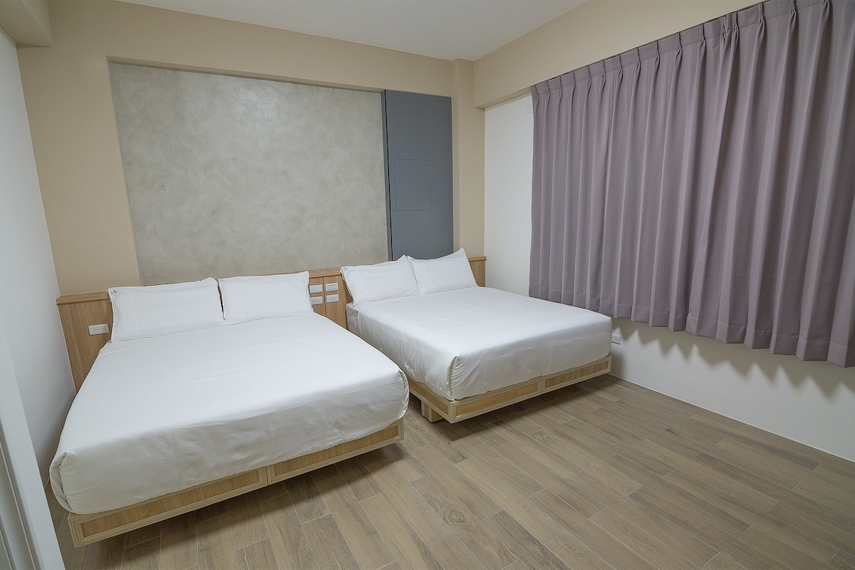 The room features two spacious beds with white linens, positioned against a light-colored wall. A large window is adorned with soft purple curtains, allowing natural light to fill the space. Smooth wooden flooring adds warmth to the overall decor.