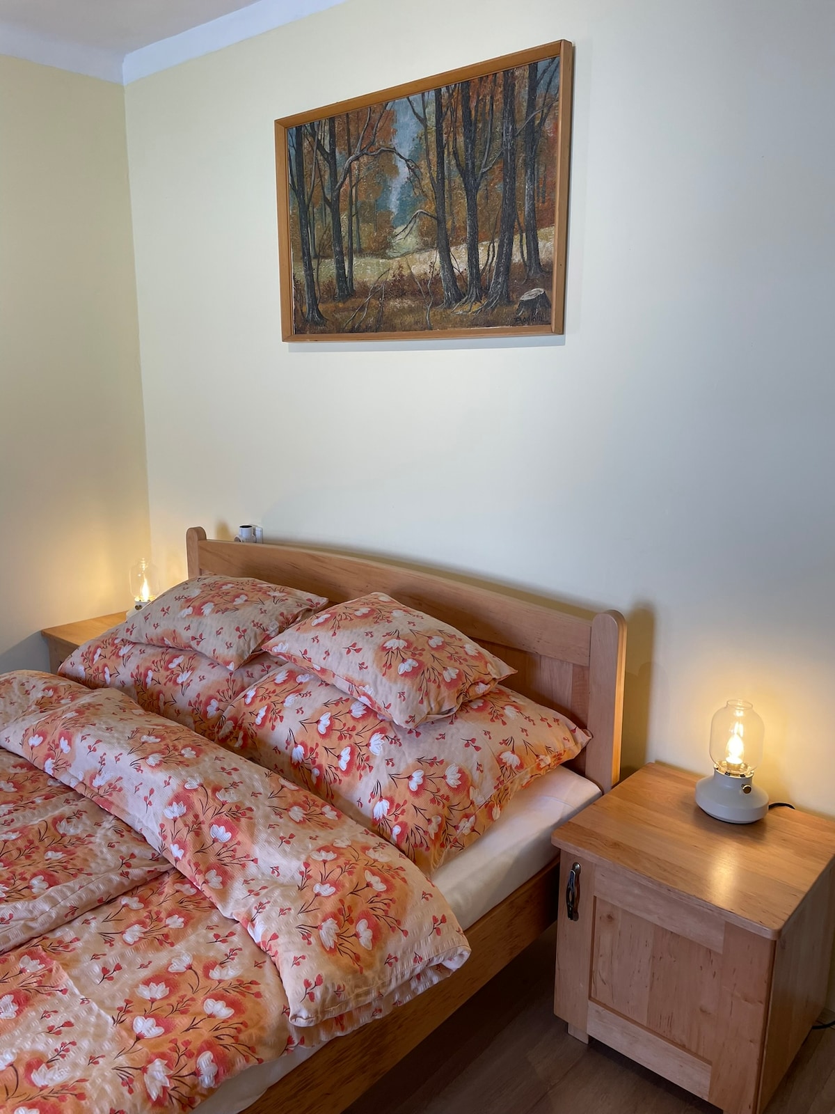 A cozy sleeping area features a wooden bed adorned with plush bedding in floral patterns. A bedside table with a warm light and a nature-themed painting hang on the pale yellow wall, contributing to a restful atmosphere.