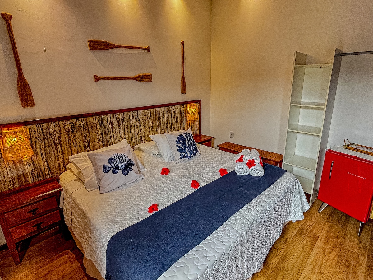 The sleeping area features a spacious bed dressed in white linens, accented by decorative pillows. Wooden paddles are displayed on the wall, with soft lighting from bedside lamps. A minimalistic shelving unit and a red cabinet provide functional storage, complemented by warm wood flooring.