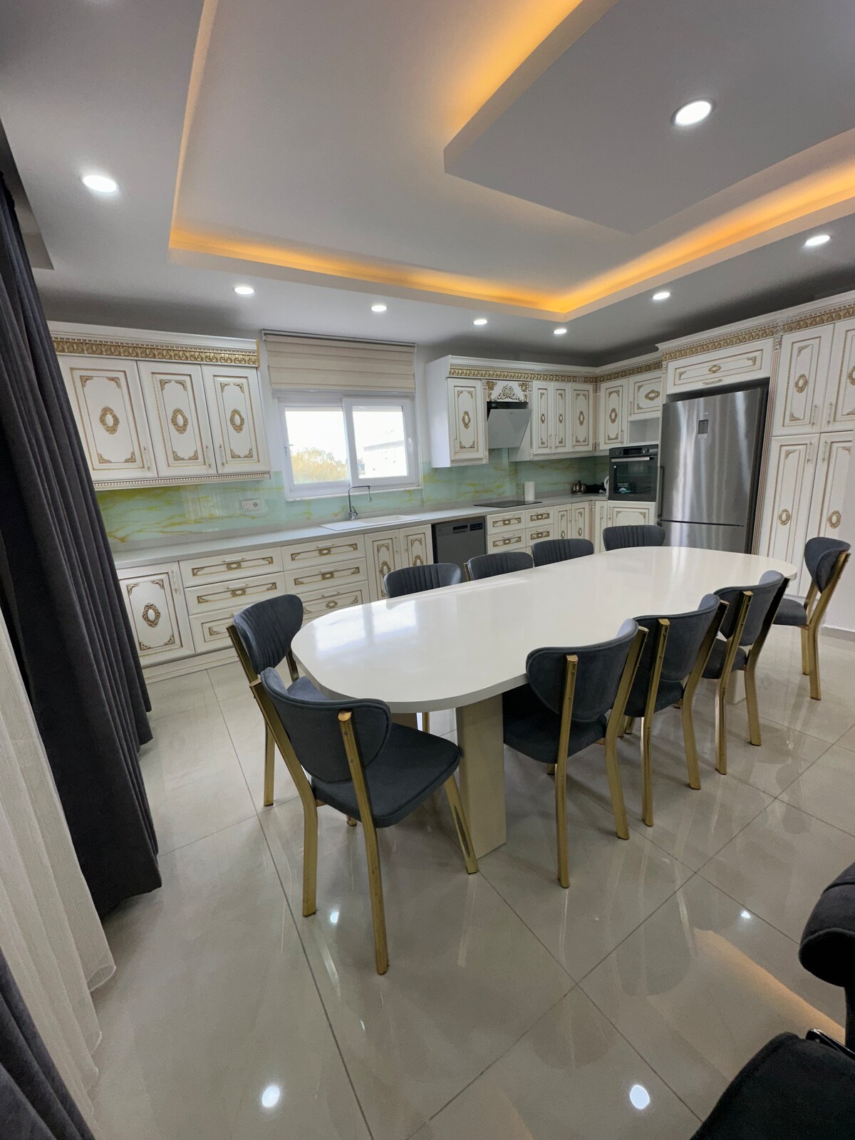 The modern kitchen features an elegant white dining table surrounded by gold-accented chairs. Well-organized cabinets display decorative elements, while high-end appliances are integrated into the design. Natural light enters through a window, and recessed lighting creates a bright atmosphere.