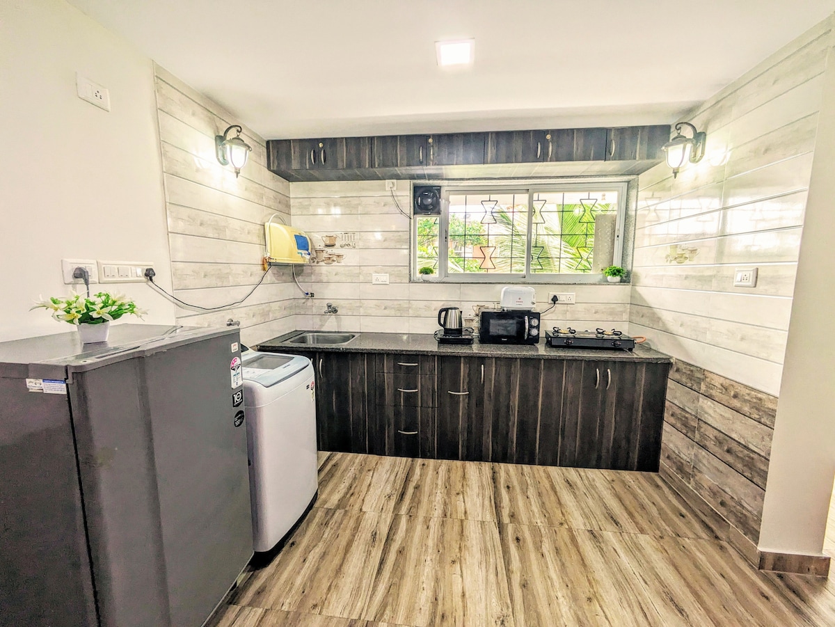 A modern kitchen features sleek wooden cabinets and a spacious countertop. A refrigerator, washing machine, and electric kettle are visible, alongside a two-burner gas stove. Natural light enters through a window, illuminating the neutral color palette of the space.